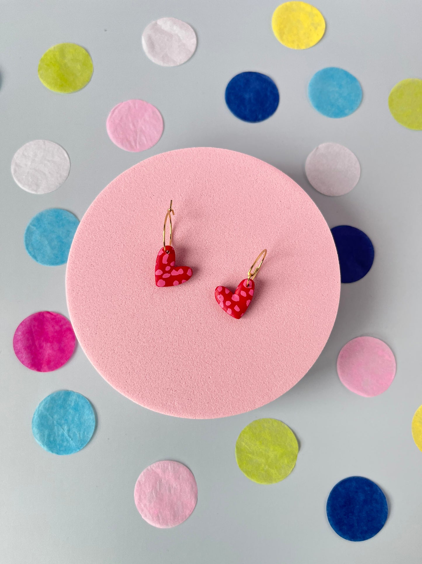 Red heart-shaped earrings on a pink stand with colorful circular patterns in the background