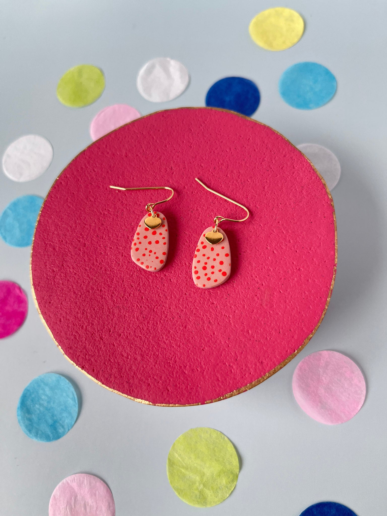 Pair of pink earrings with gold accents on a pink circular stand with colorful confetti background