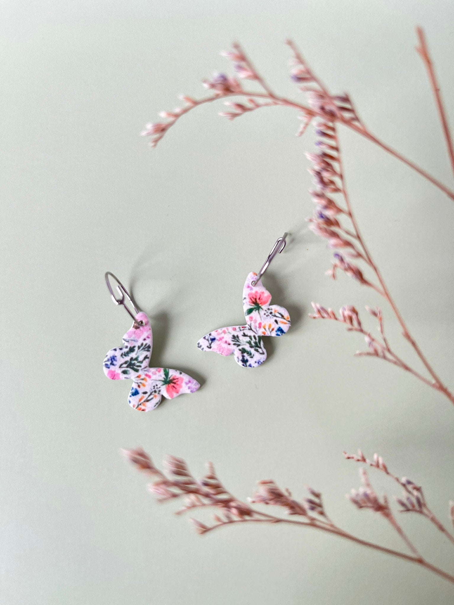Butterfly-shaped earrings with floral design on a light gray background with dried branches.
