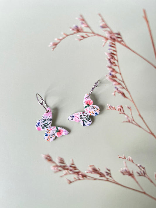 Butterfly-shaped earrings with floral design on a light gray background with dried branches.