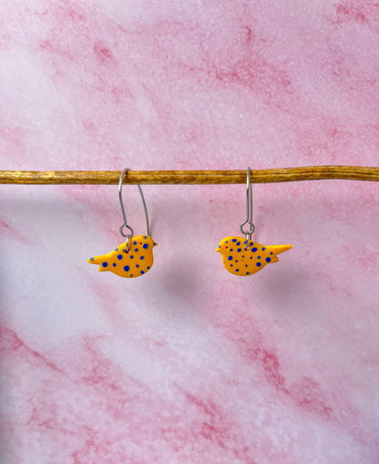 Yellow bird-shaped earrings with blue spots on a pink marble background